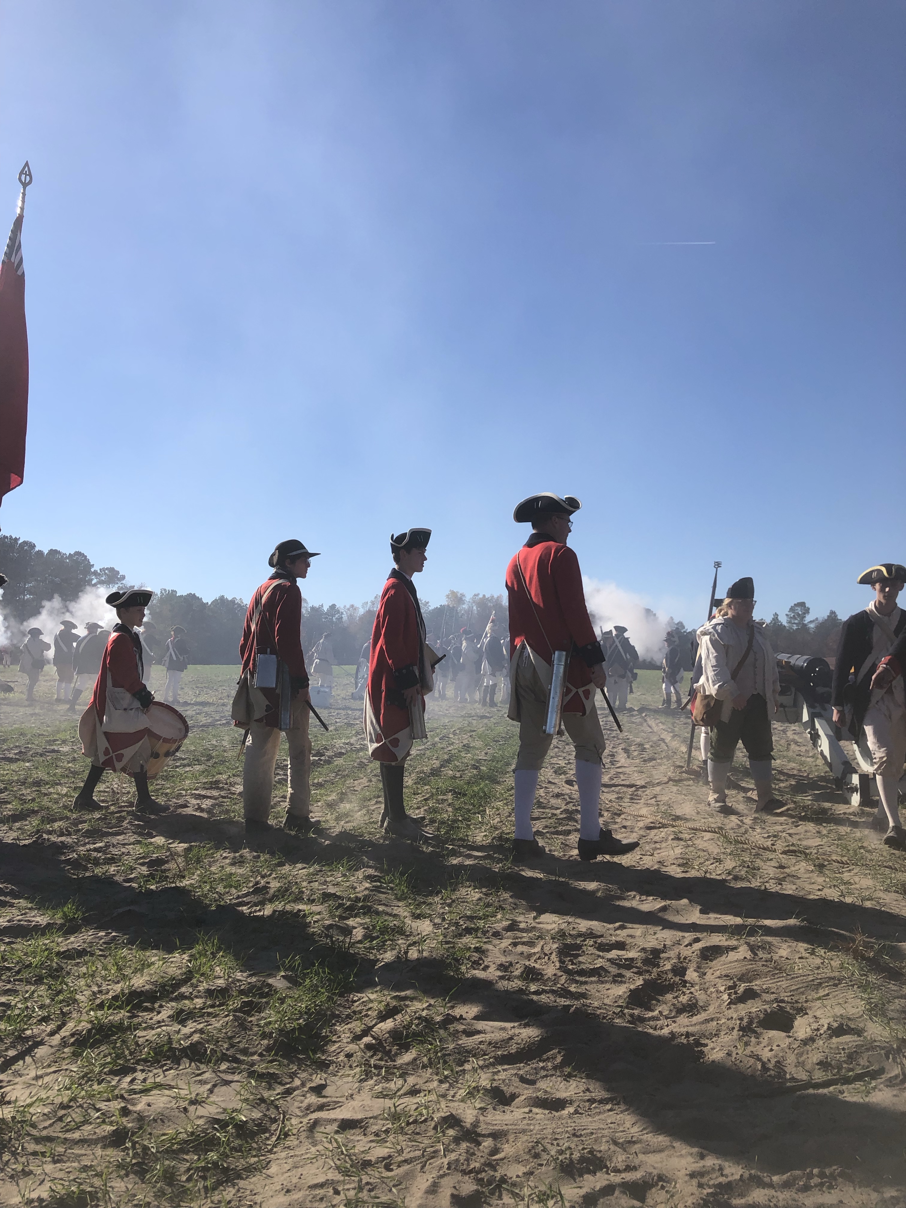 Reenactors in red coats, including a drummer, watching from the field