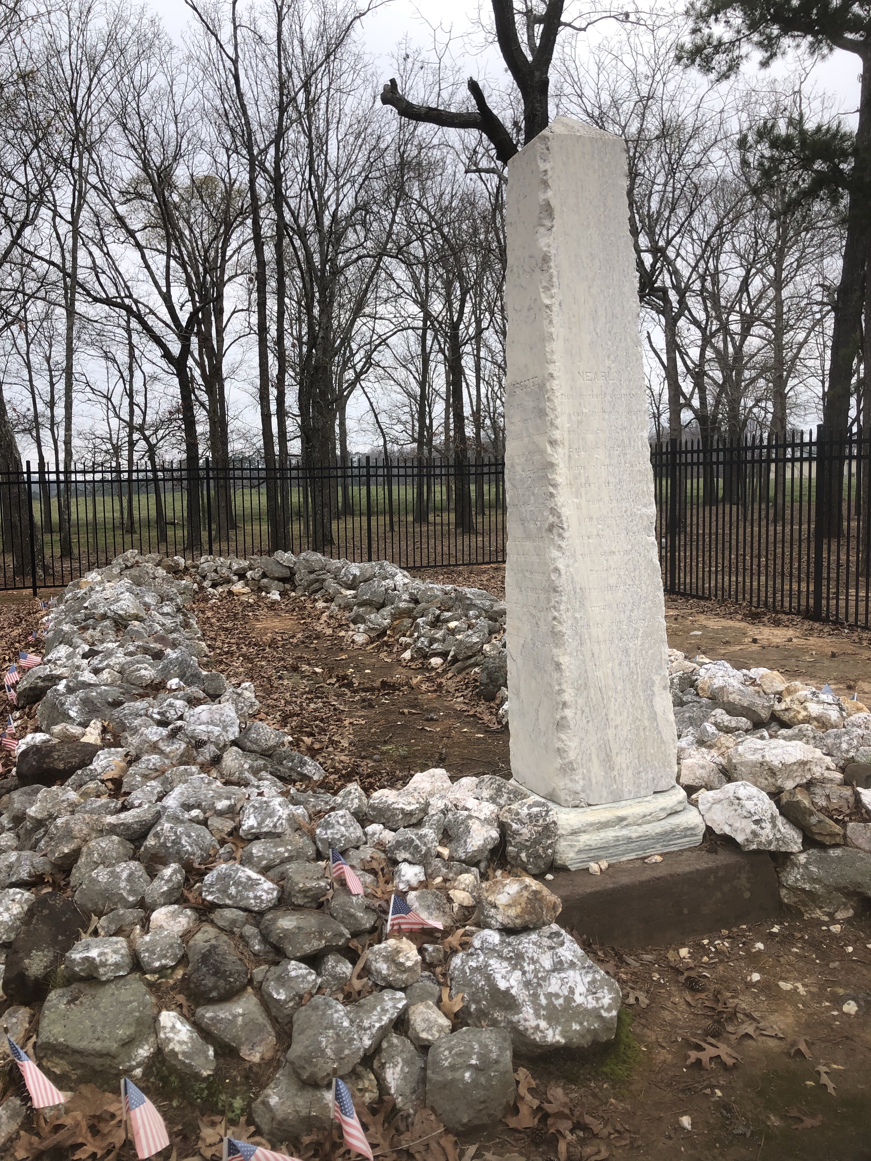 Tall white marble marker beside the stone-covered mass grave at the Waxhaws battlefield, with small American flags