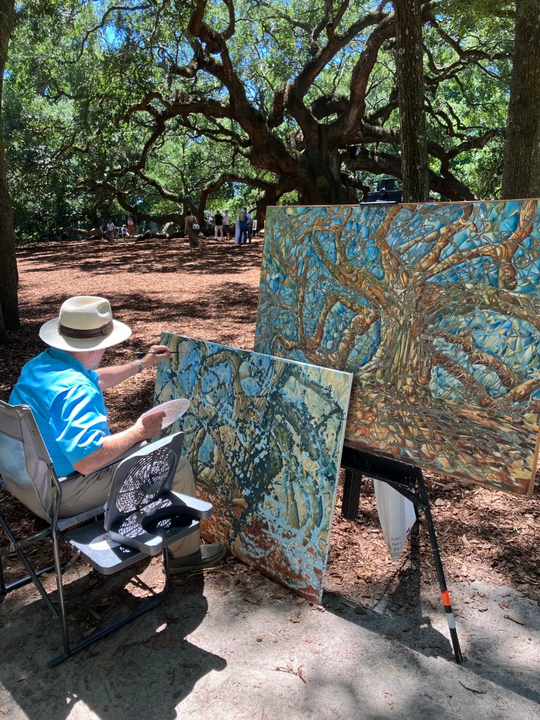 Artist in a sun hat painting the Angel Oak beside his finished canvases