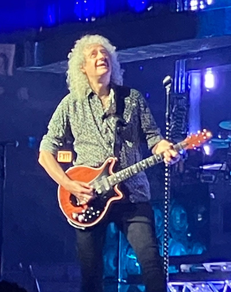 Brian May playing his red guitar under blue stage lights