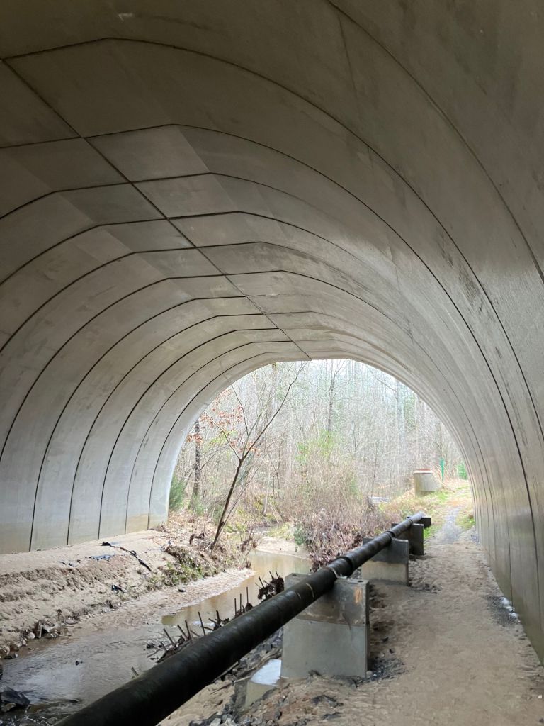 Trail running through a large metal culvert tunnel beside a pipe
