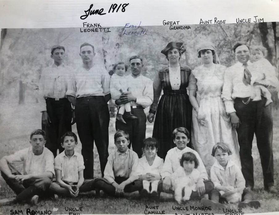 Outdoor family portrait, June 1918: a teenage boy and five adults standing, two of the men holding babies, and seven children seated on the grass in front
