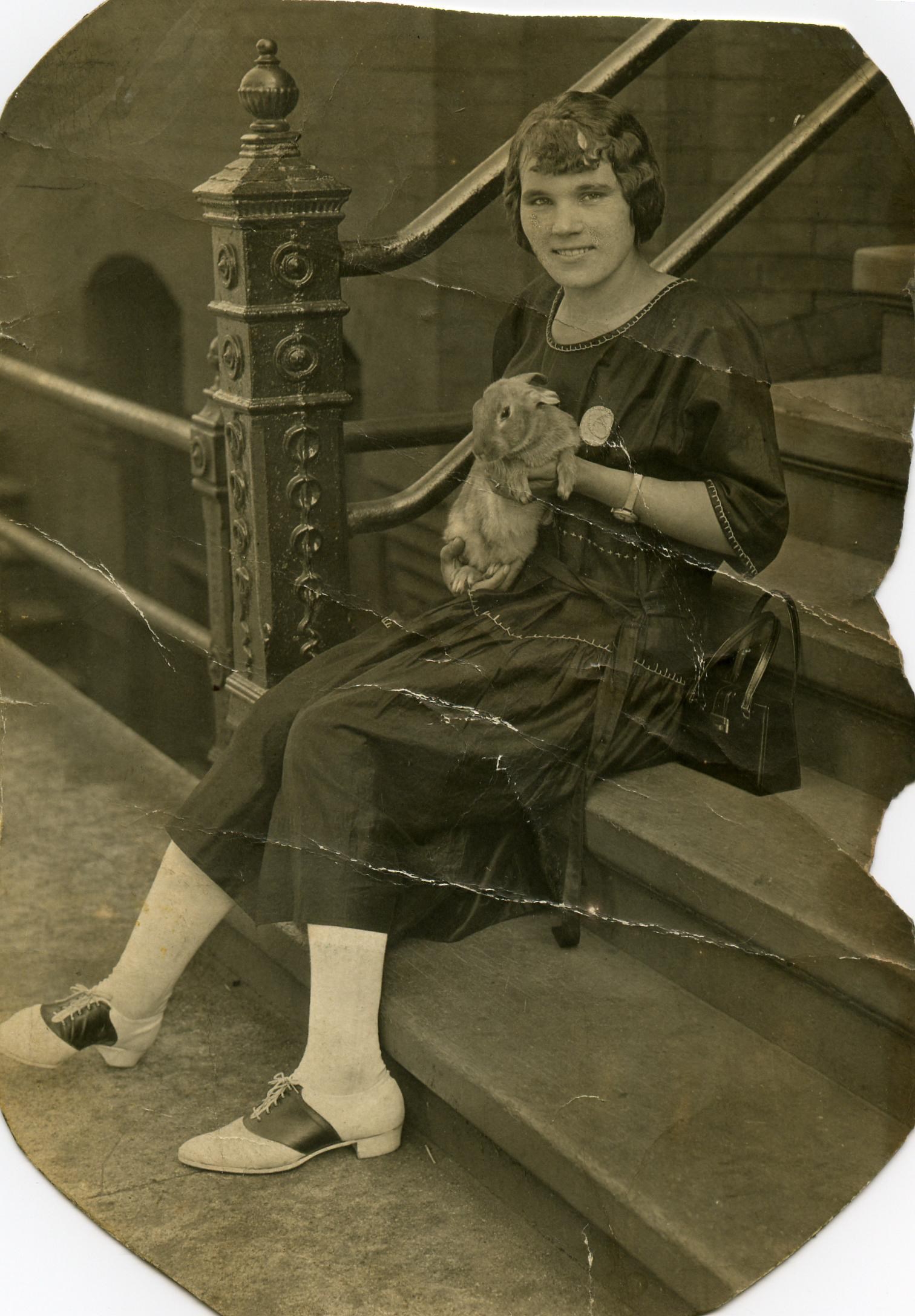 Olena, a young woman with bobbed hair, seated on stone steps beside an ornate iron newel post, holding a rabbit. She wears a dark dress with light stitching, a round pin, a wristwatch and saddle oxfords, with a handbag beside her.