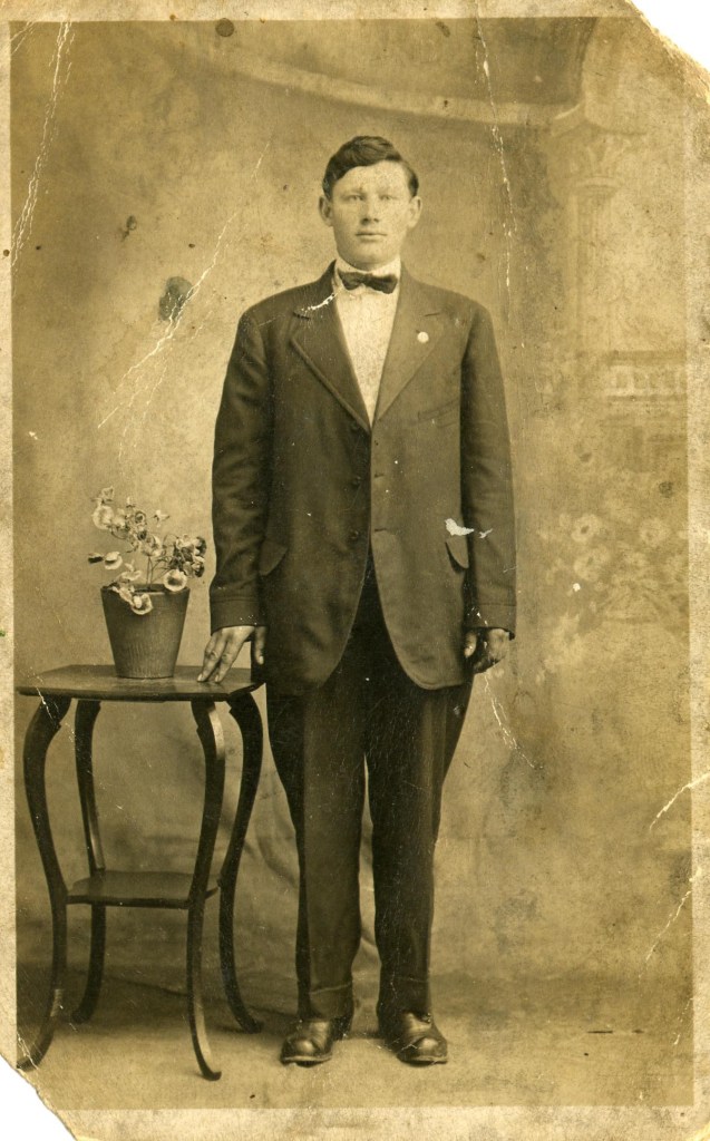 Studio portrait of Roman Patrick standing in a dark suit and bow tie beside a small table with a potted plant